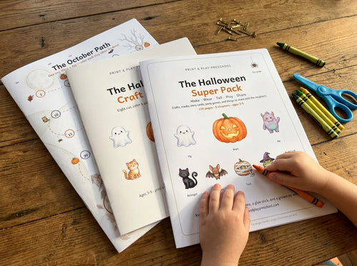 The Halloween Super Bundle: the three printed sets, The Halloween Super Pack, The Halloween Craft Album and The October Path, fanned on a kitchen table with a child's hand holding a crayon
