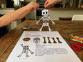 The Halloween Super Pack: the Dancing Bones puppet from Craft 1, cut out, joined with paper fasteners and held up by a child's hands next to the printed page