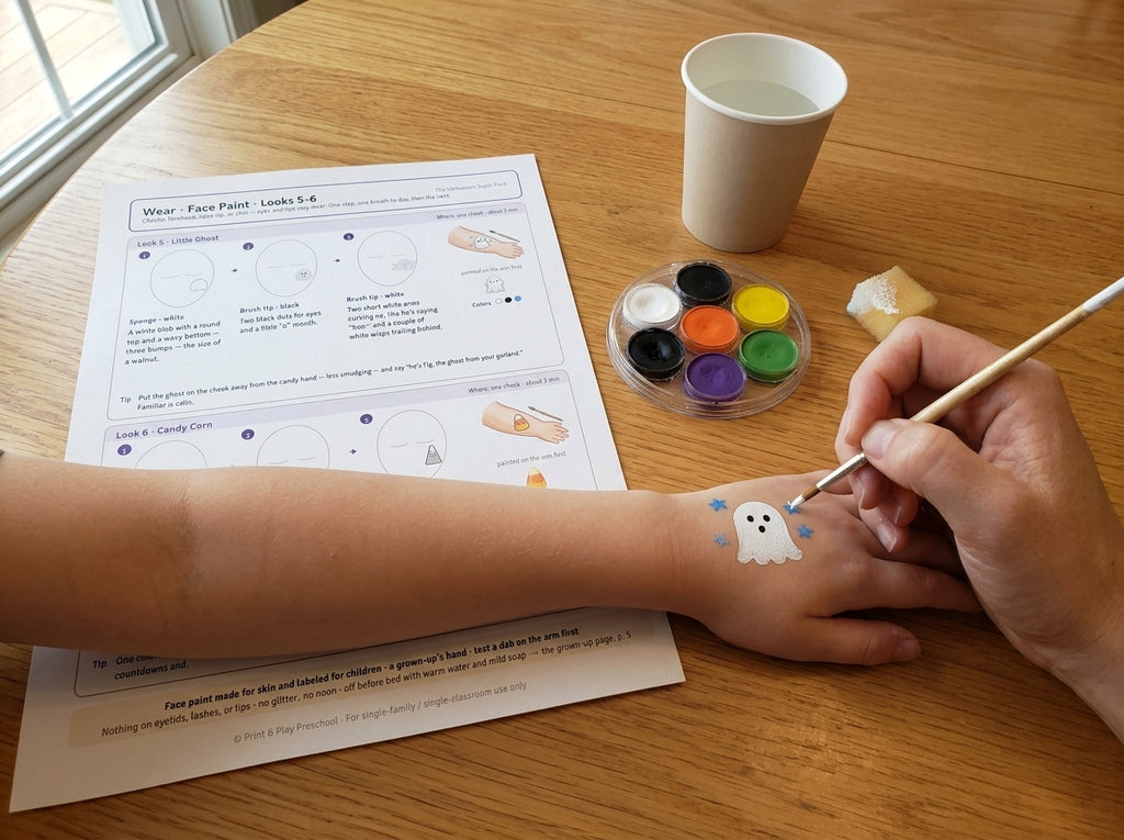 The Halloween Super Pack: Face Paint Look 5, the Little Ghost, painted on a child's forearm by a grown-up's hand, with the printed face-paint page, a palette and a brush on the table