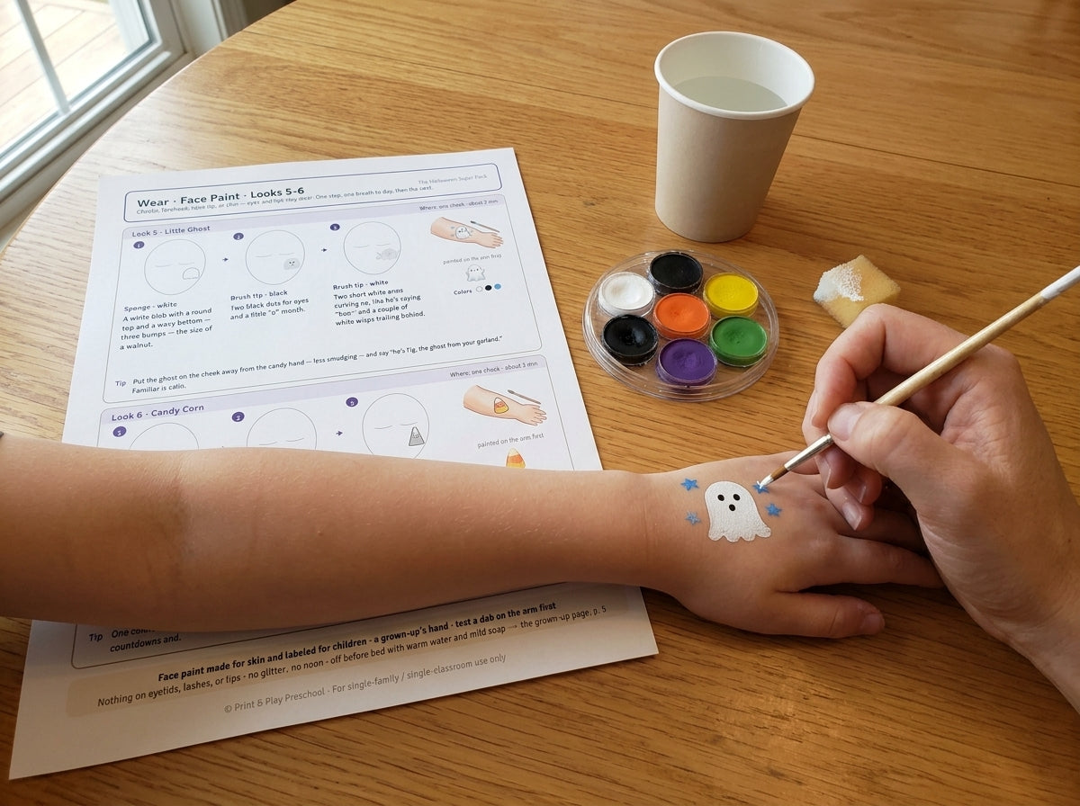 The Halloween Super Pack: Face Paint Look 5, the Little Ghost, painted on a child's forearm by a grown-up's hand, with the printed face-paint page, a palette and a brush on the table